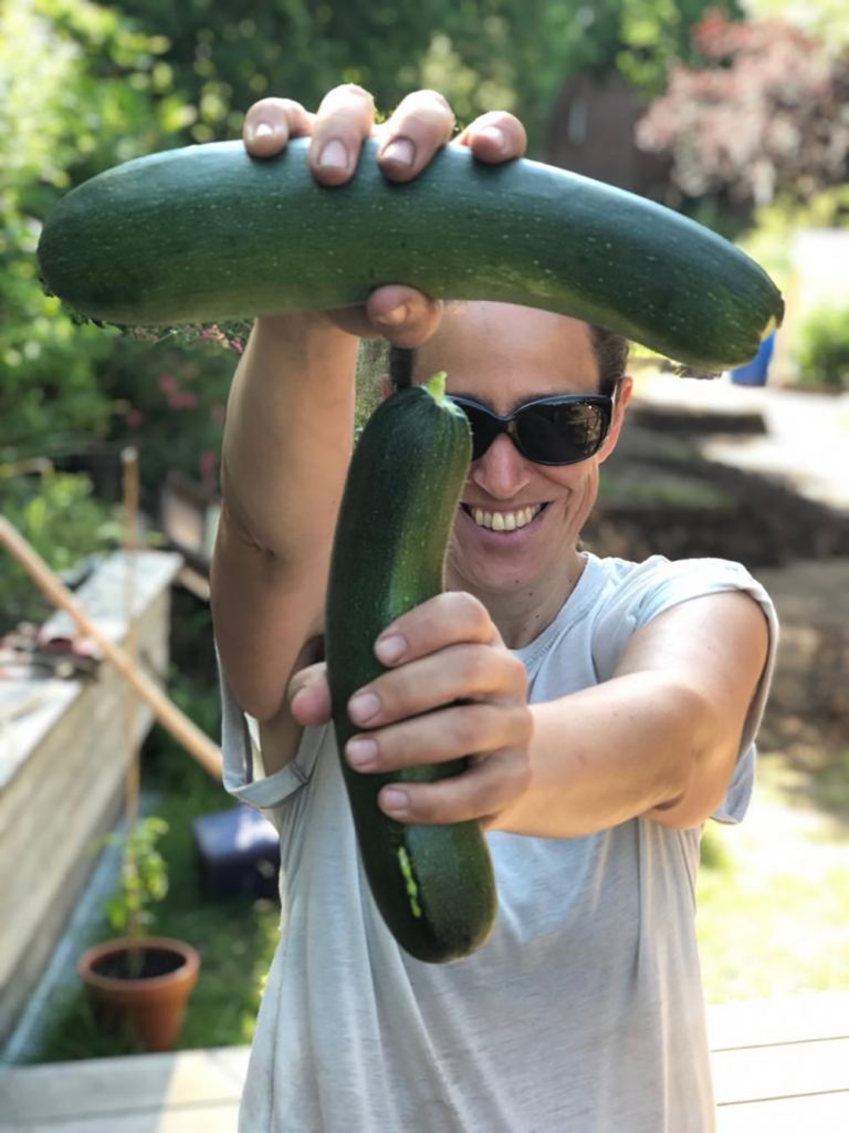 Eine Person mit Sonnenbrille und weißem Hemd steht im Freien, lächelt und hält zwei große Zucchinis vor ihr Gesicht. Eine Zucchini hält sie waagerecht über dem Kopf, die andere senkrecht vor der Brust. Im Hintergrund sind Grünpflanzen von Urban Gardening Hamburg zu sehen.